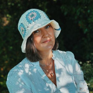 Könnte beinhalten: Ein cremefarbener Häkel-Eimerhut mit einem Blumenmuster in Hellblau und Rosa. Der Hut hat eine breite Krempe und ein blaues Akzentband. Ein modisches Accessoire zum Schutz vor der Sonne.