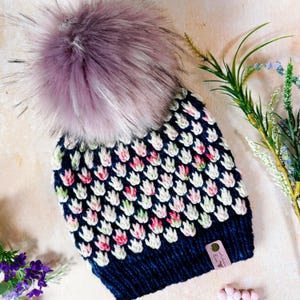 May include: A navy blue knitted hat with a white and floral pattern, topped with a large, fluffy, lavender-coloured pom-pom. The hat has a small tag attached. The hat is a winter accessory.