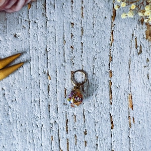 May include: A small, clear glass charm filled with colorful beads and glitter, suspended from a gold-colored ring. The charm is round and the beads are various sizes and colors. The background is a weathered, light blue wooden surface.