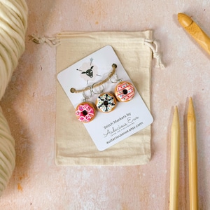 May include: Set of three donut stitch markers with pink, black and white frosting. The markers are on a white card with the text "Stitch Markers by Audacious Eve Leave the Frowns Behind audaciousweve.etsy.com"