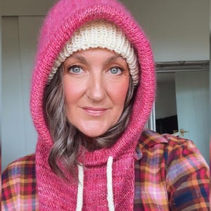 May include: A knitted hood in a vibrant pink hue, paired with a cream-colored beanie. The hood features a drawstring and is worn over a plaid shirt. The overall look is cozy and stylish, perfect for cold weather.