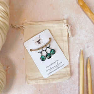 May include: Set of three green and white stitch markers with a floral design. The markers are on a white card with the text "Stitch Markers by Audacious Ewe" and "Leaves the Flock Behind" and a website address. The card is in a small white drawstring bag.