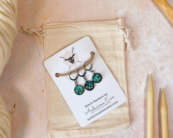 May include: Set of three green and white stitch markers with a floral design. The markers are on a white card with the text "Stitch Markers by Audacious Ewe" and "Leaves the Flock Behind" and a website address. The card is in a small white drawstring bag.
