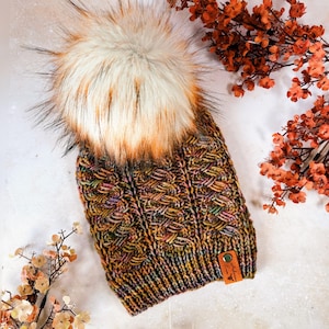 May include: A hand-knitted beanie with a large, fluffy faux fur pom-pom. The hat features a textured cable knit pattern in shades of brown, gold, and green. A small leather tag is sewn on the side. The pom-pom is cream, brown, and orange.