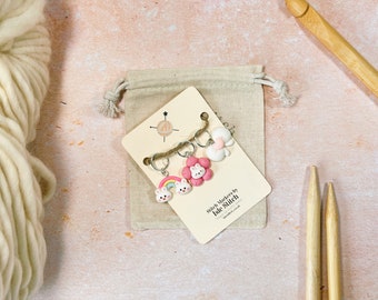 May include: A set of four colorful stitch markers with a rainbow, a flower, a bunny, and a cloud design. The markers are packaged in a small burlap bag and come with a card that says "Stitch Markers by Isle Stitch Handmade in the UK".