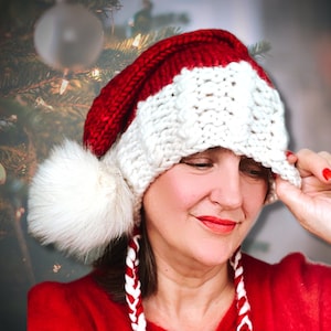 Könnte beinhalten: Eine weiße und rote gestrickte Weihnachtsmütze mit einem weißen Bommel und einer weißen und roten geflochtenen Schnur. Die Mütze wird von einer Person mit braunen Haaren getragen.
