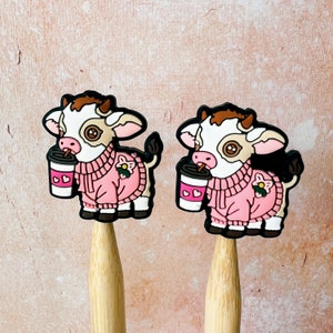 May include: Two pink cow-shaped pencil toppers with brown spots and white faces. Each cow is holding a pink and white cup with a heart design. The cows are wearing pink sweaters with a flower design.