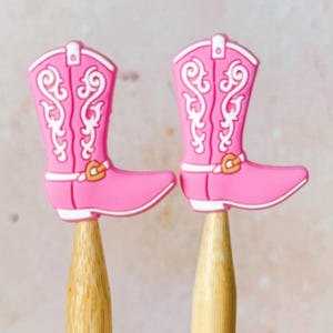 May include: Two pink cowboy boot-shaped toppers with white scrollwork and gold-coloured buckles. The boots are mounted on wooden sticks. The toppers are a fun, decorative item.