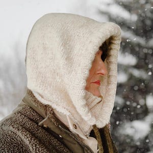 May include: A cream-colored knitted hood, worn over a jacket, provides warmth. The hood is made of thick yarn and has a textured appearance. The person wearing the hood is seen in a snowy outdoor setting, suggesting cold weather.