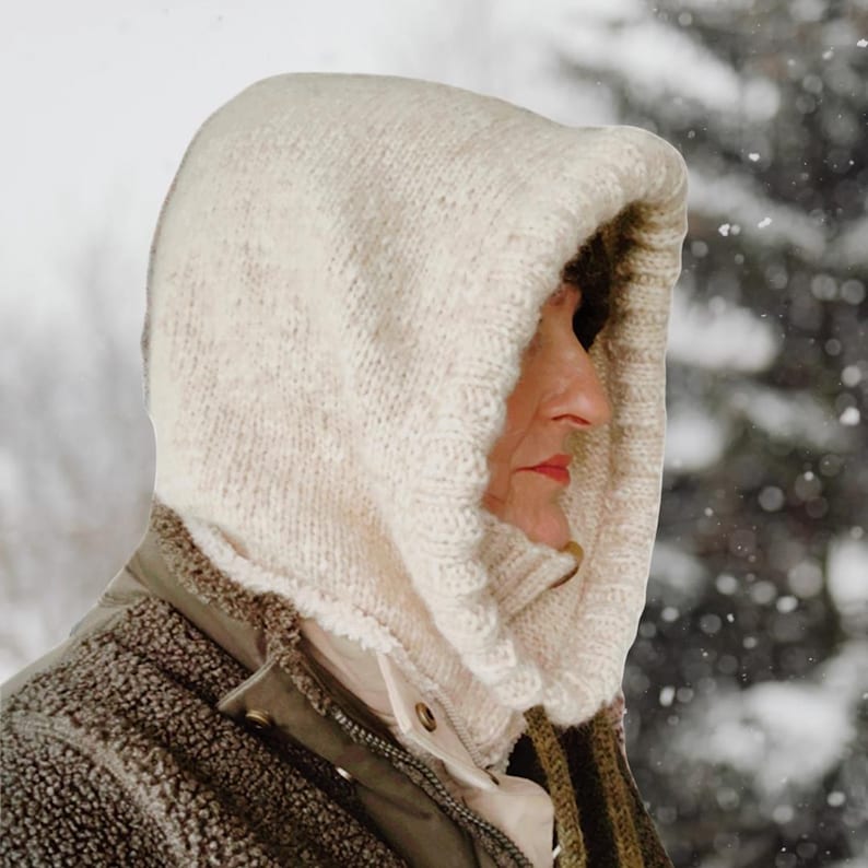 May include: A cream-colored knitted hood, worn over a jacket, provides warmth. The hood is made of thick yarn and has a textured appearance. The person wearing the hood is seen in a snowy outdoor setting, suggesting cold weather.