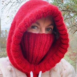 May include: A vibrant red knitted hood with a built-in face covering. The hood is thick and textured, with a ribbed knit design on the face covering. White drawstrings are visible at the neck. The person is wearing a cream-colored sweater.