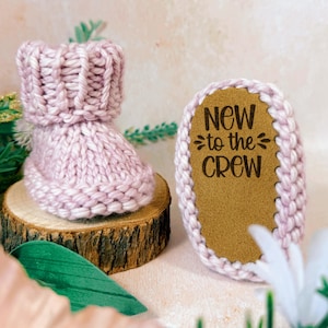 May include: A knitted baby bootie and a matching sole, both in a soft pink hue. The sole features the text "NEW to the CREW" in a dark brown color. The bootie is displayed on a small wooden slice, suggesting a baby-themed product.