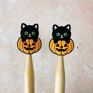 May include: Two Halloween-themed pencil toppers. Each features a black cat with green eyes perched atop an orange jack-o'-lantern. The pencils are made of light-colored wood. The background is a light, textured surface.