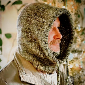 May include: A knitted hood in shades of olive green and brown, with a drawstring closure. The hood is made of a textured yarn and is worn over a jacket. The overall style is casual and warm, suitable for cold weather.