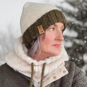 May include: A knitted winter hat with a cream-colored crown and an olive green brim. A small leather tag is sewn on the side. The hat is worn with a cream-colored neck warmer and a green jacket.