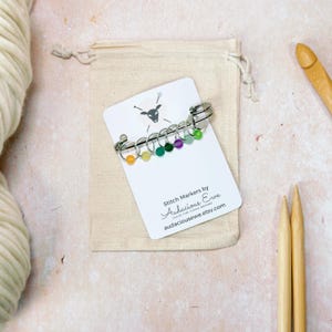 May include: A set of colorful stitch markers with a silver ring and a small bead. The markers are arranged in a rainbow pattern. The card reads "Stitch Markers by Audacious Ewe Leave the Flock Behind audaciousewe.etsy.com"