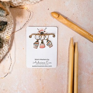 May include: A set of three stitch markers with winter-themed designs, including mittens and a bell, attached to a card with the text "Stitch Markers by Audacious Ewe". A wooden crochet hook and two knitting needles are also present.