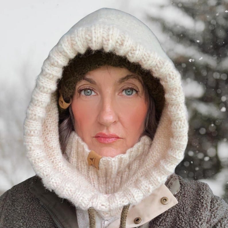 May include: Cream-colored knitted hood with a ribbed cowl neck and wooden toggle closures. The hood is worn over a dark green hat and a gray jacket. The background shows a snowy, blurred winter scene.