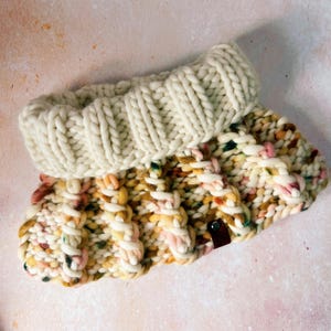 Könnte beinhalten: Ein weißer und mehrfarbiger gestrickter Halsschmuck mit einem braunen Lederanhänger. Der Halsschmuck ist aus dickem Garn gefertigt und hat ein Zopfmuster.