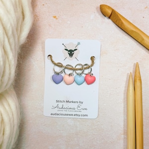 May include: Set of four heart-shaped stitch markers in lavender, pink, blue, and coral colors. The markers are strung on a silver chain and are displayed on a white card with the text "Stitch Markers by Audacious Ewe Leave the Flock Behind audaciousewe.etsy.com".