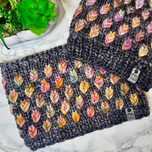 Könnte beinhalten: Eine graue Strickmütze und ein passender Schal mit einem bunten Blättermuster. Mütze und Schal sind aus einem dicken Garn gefertigt und haben ein weiches, gemütliches Gefühl. Die Mütze hat ein kleines Etikett mit der Aufschrift "Two Little Hooligans".