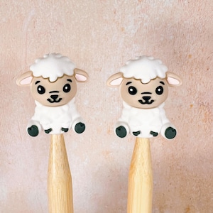 May include: Two wooden knitting needles with white sheep-shaped silicone toppers. The sheep have black eyes and a brown nose.