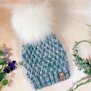 Puede incluir: Un gorro de punto azul y morado con un pompón de piel sintética blanca. El gorro tiene un patrón de punto texturizado y una etiqueta de cuero marrón con el texto "Stitch & Love" en él.