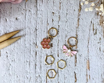 May include: Four gold stitch markers with a pink honeycomb design and a pink bee design. The stitch markers are on a white wooden surface.