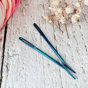May include: Two blue metal needles with a pointed tip and a large eye for threading. The needles are lying on a white wooden surface.