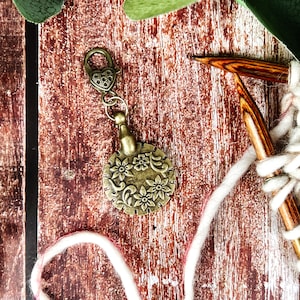 May include: A bronze stitch marker featuring a floral pattern and a heart-shaped clasp. The marker is connected to white and pink yarn, with wooden knitting needles visible. The background is a wooden surface.