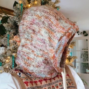May include: A hand-knitted hood in a mix of colors, including orange, pink, and cream. The hood is part of a sweater, with a patterned vest visible below. A white drawstring is visible on the side of the hood. The background includes a decorated Christmas tree.