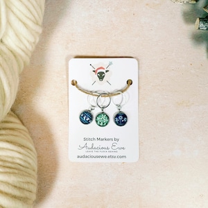 May include: A set of three stitch markers with decorative designs, attached to a white card with a jute string. The markers have silver-colored rings and feature blue and green patterns. The card includes the text "Stitch Markers by Audacious Ewe."