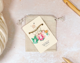 May include: Set of three stitch markers featuring a green watering can, a pink flower, and a white bunny with an orange flower. The markers are packaged on a card with the text "Stitch Markers by Isle Stitch" and a website address.