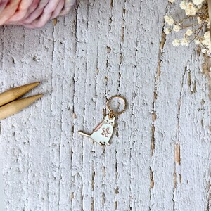 May include: White cat stitch marker with a pink flower on its chest. The marker is made of metal and has a gold ring.