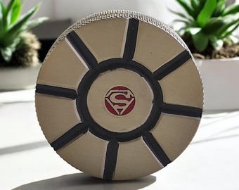 May include: A round, silver-colored puck with a black design and a red Superman logo in the center.