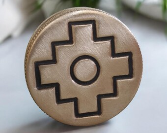 May include: A circular, bronze-coloured object with a raised, geometric design. The design features a cross-like shape with a central circle. The object has a textured surface and a circular edge.