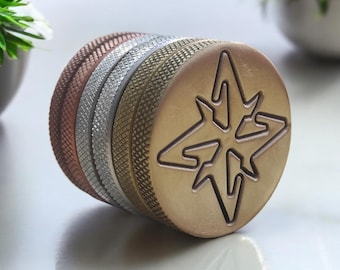 May include: A set of four round, metal, decorative stamp heads. The stamp heads are stacked on top of each other. The top stamp head is gold and features a starburst design. The other stamp heads are silver, copper, and gold.