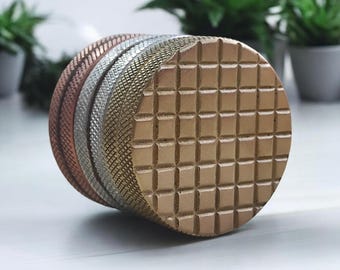 May include: A stack of four round, textured metal discs. The top disc is gold and has a grid pattern. The second disc is silver and has a textured pattern. The third disc is copper and has a textured pattern. The bottom disc is copper and has a smooth surface.