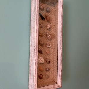 May include: A white wooden frame with a glass front displays a collection of seashells on a woven background.