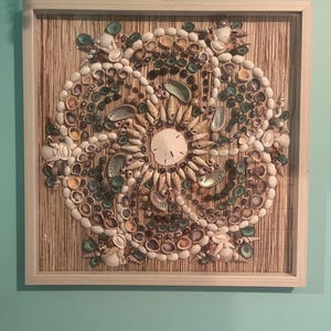 May include: A framed artwork featuring a circular design made of various seashells. The shells are arranged on a textured, light brown background. The shells are in shades of white, tan, and green, with a sand dollar in the center.