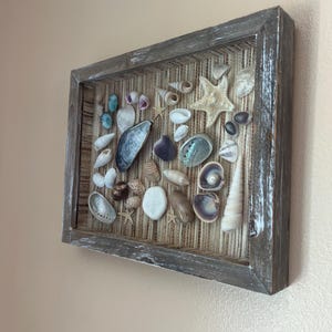 May include: A rectangular shadow box frame with a distressed dark wood finish containing a collection of various seashells, including a starfish, a sand dollar, and an iridescent abalone shell, arranged on a textured tan woven fabric backing.