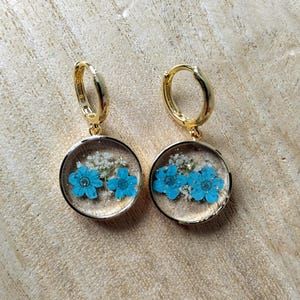 Peut inclure: Boucles d'oreilles créoles dorées avec un pendentif rond présentant des fleurs bleues et des fleurs séchées blanches enchâssées dans de la résine transparente.