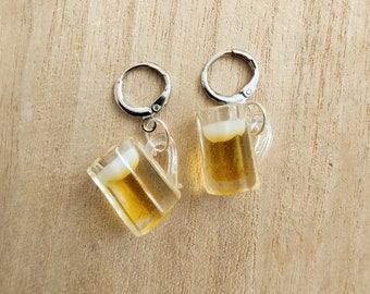 May include: A pair of novelty earrings featuring miniature beer mugs. Each clear plastic mug is filled with a golden liquid and topped with a white foam. The earrings have silver-colored lever-back closures and are set against a wooden background.
