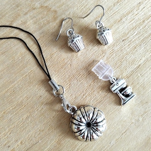 May include: A pair of silver-toned cupcake earrings with hook backings, a silver-toned bundt cake charm on a black cord strap, and a silver-toned stand mixer charm with a clear plastic plug attachment for electronic devices.