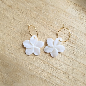 Peut inclure: Une paire de boucles d'oreilles créoles dorées avec des breloques de fleurs blanches. Les fleurs sont faites d'un matériau blanc qui ressemble à de la nacre.