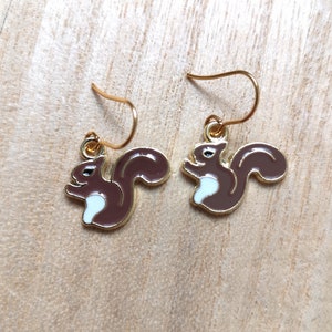 Peut inclure: Une paire de boucles d'oreilles dorées avec un motif d'écureuil brun et un ventre blanc. Les écureuils sont tournés dans des directions opposées.