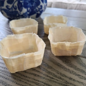 May include: Four small, square, beige paper baskets are arranged in a group. The baskets have a textured appearance and are open at the top. A blue and white patterned ceramic bowl is visible in the background.