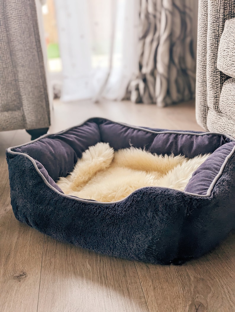 May include: A dark blue pet bed with a plush, soft lining and a cream-colored faux fur insert. The bed is sitting on a light brown wood floor.