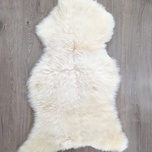 May include: A white sheepskin rug with a soft, fluffy texture. The rug is laid out flat on a wooden floor.