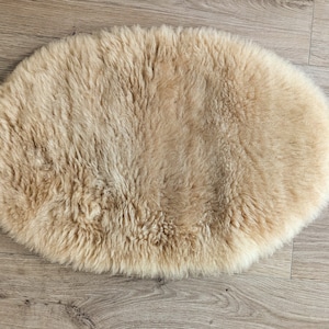 May include: A light brown sheepskin rug with a soft, fluffy texture. The rug is oval shaped and is laying flat on a wooden floor.
